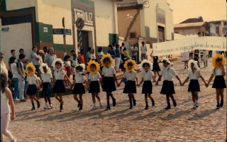 'Estudantes em uniforme e fantasia de flor durante desfile cívico, vendo-se o barracão da Tammuz Década de 80