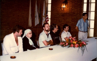 'João Bosco, Madre Emanuelle Favale, prefeito Geralo Barbosa Leão e outros indivíduos em mesa durante reunião no Clube Social, com um homem de pé discursando Década 80