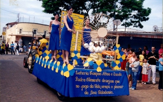'Carro alegorico da Loteria Campos Altos cin Bruna, Júlia e outra moça, em desfile pela Praça Benedito Valadares, vendo-se ao fundo o bar do Osvaldo e o treiler Água na Boca ano 2000