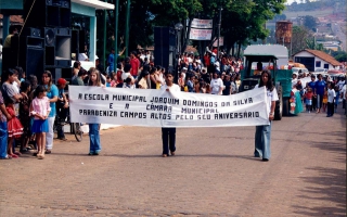 'Estudantes da Escola Municipal Joaquim Domingos da Silva em desfile pela Praça Benedito Valadares, assistidos por multidão, e carregando faixa ano 2000