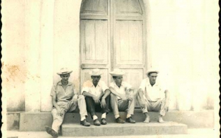 'Quatro homens usando chapéu sentados à porta de uma edificação que parece ser uma igreja Década de 50