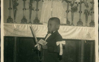 'Criança não identificada em frente ao altar da Igreja Matriz de Santa Terezinha segurando nas mãos vela e terço, no braço esquerdo laço de fita branca década de 80