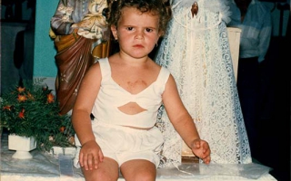 'Criança não identificada sentada em cima da mesa ao lado de imagens sagradas na Sala dos Milagres no Segundo Santuário de Nossa Senhora Aparecida década de 90