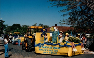 Desfile de carros alegóricos durante comemoração do cinquentenário da emancipação do município, vendo-se em primeiro plano um carro alegórico alusivo ao Banco do Brasil, com meninas segurando flores, seguindo outros carros alegóricos pela rua. 1994