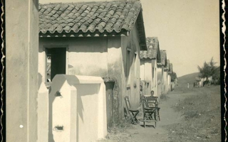 'Residências em estilo colonial em uma rua de terra, com cadeiras na frente de uma das casas e ao fundo crianças não identificadas