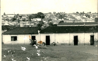 'Primeiro prédio da Escola Elizena Leão, ao fundo mais a esquerda a Igreja Matriz de Santa Terezinha na porta e na janela pessoas não identificada década de 60