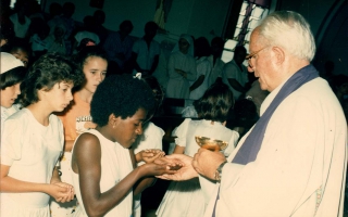 'Criança recebendo a Primeira Comunhão, no lado direito Padre Marques e no lado esquerdo ao fundo com chapéu branco Nair Caetano  década de 80